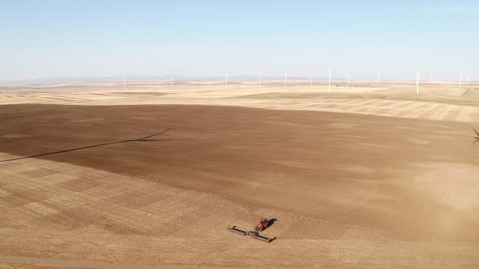 Aerial view of a red implement working across brown stubble field with wind turbines on horizon