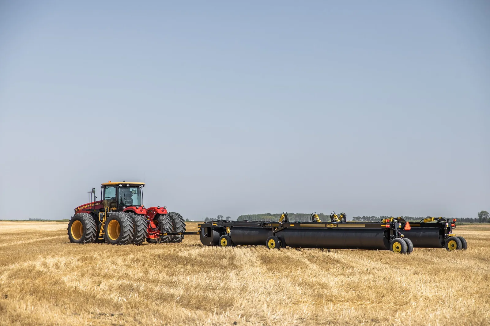 Red and black land roller with dual wheels behind yellow tractor in harvested grain field